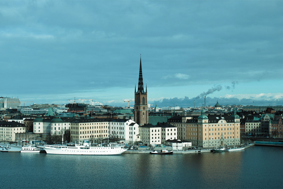 A scenic cityscape of Stockholm with a tall church spire rising above a harbor lined with boats and buildings under a cloudy blue sky.
